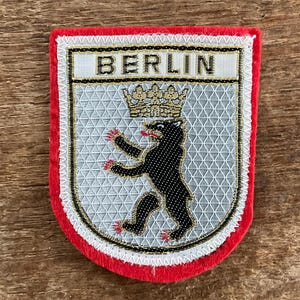 Puede incluir: Un parche de Berlín con borde rojo. El parche presenta la palabra "BERLIN" sobre un oso negro con corona, sobre un fondo plateado con un patrón geométrico. El oso tiene garras rojas.