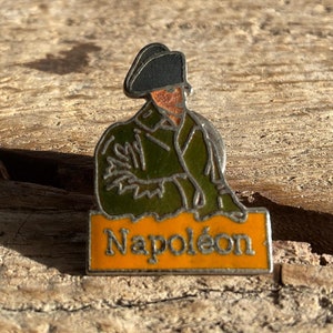 Op de afbeelding: Een vintage emaille speld met een afbeelding van Napoleon Bonaparte in een groene jas en zwarte hoed. De speld heeft een zilveren rand en een oranje rechthoekige basis met de naam "Napoléon" in gouden letters. Geplaatst op een rustiek houten oppervlak.
