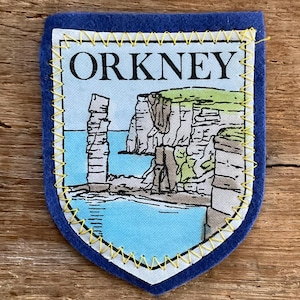 May include: A blue felt patch featuring a printed image of the Orkney Islands. The image shows a coastal scene with cliffs, sea stacks, and the word "ORKNEY". The patch is edged with yellow stitching.