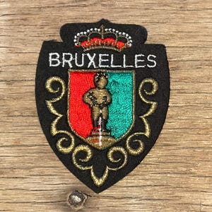 Op de afbeelding: Een zwarte geborduurde patch met een schildontwerp en de tekst BRUXELLES in het wit. Het schild heeft een rode en groene achtergrond met een gouden figuur in het midden, bekroond door een kroon met rode accenten en gouden krulwerk.