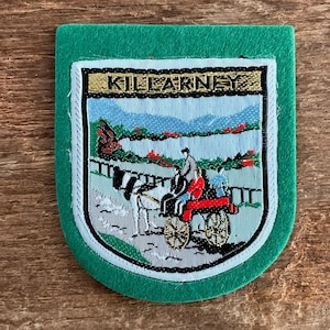 Op de afbeelding: Een groene vilt patch met een geborduurd beeld van een paard en wagen voor een meer en bergen. Het woord "KILLARNEY" is bovenaan de patch geborduurd.