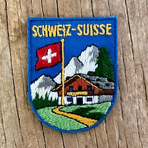 Könnte beinhalten: Ein rechteckiger bestickter Aufnäher mit blauem Hintergrund und dem gelben Schriftzug SCHWEIZ-SUISSE. Das Motiv zeigt eine rote Schweizer Flagge, ein Holzchalet, grüne Tannen, einen gelben Weg und weiße schneebedeckte Berge im Hintergrund.