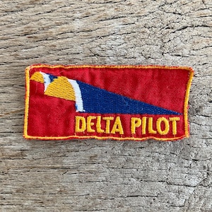 Op de afbeelding: Een rechthoekige patch met een rode achtergrond en een gele rand. De patch heeft een blauw en wit ontwerp met een gele vogel en de woorden "DELTA PILOT" in het geel.