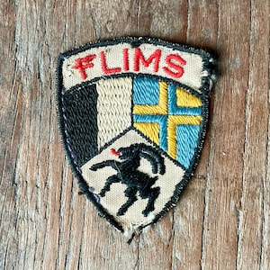 Op de afbeelding: Geborduurde patch met het woord "FLIMS" in het rood. De patch heeft een schildontwerp met een zwart, wit en blauw kleurenschema en een zwart silhouet van een springend dier. De patch ligt op een houten oppervlak.