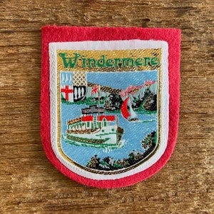 Op de afbeelding: Geborduurde patch met een rode rand en een schildvormig ontwerp. De patch toont het woord "Windermere" in gouden letters, een boot op een meer en andere landschappelijke elementen. De achtergrond is een houten oppervlak.