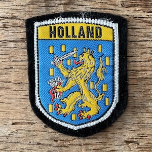 Puede incluir: Un parche en forma de escudo con la palabra "HOLLAND" en amarillo sobre un fondo azul. El parche presenta un león amarillo sosteniendo una espada. El parche está bordeado en blanco y negro.