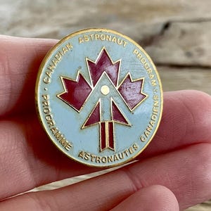 Op de afbeelding: Een ronde speld met een lichtblauwe achtergrond en gouden rand. De speld heeft een rood esdoornbladontwerp en de gouden tekst "CANADIAN ASTRONAUT PROGRAMME". Een verzamelobject.