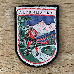 Könnte beinhalten: Rechteckiger Aufnäher mit dem Wort "ALTENMARKT" oben. Der Aufnäher zeigt einen Wanderer in roter Kleidung, eine Bergkette und einen Fluss. Der Aufnäher ist von einem weiß-roten Nahtmuster und einem schwarzen Rand umgeben.