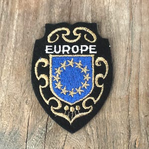 May include: A shield-shaped embroidered patch with a black background and gold decorative scrollwork. The word EUROPE is stitched in white above a blue central shield containing a circle of twelve gold stars, mimicking the European Union flag design.