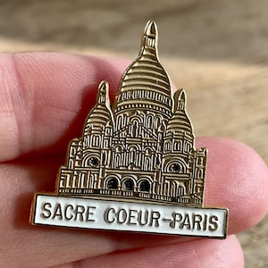 Op de afbeelding: Een goudkleurige metalen pin met de Sacré-Cœur Basiliek in Parijs. De pin heeft een witte rechthoekige basis met de woorden "SACRE COEUR-PARIS" in zwarte letters. De pin wordt in een hand gehouden.