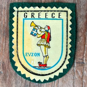 Puede incluir: Un parche de recuerdo vintage de Grecia con un borde verde. El parche presenta un diseño de escudo con la palabra "GREECE" en la parte superior y "EVZON" en la parte inferior. Dentro del escudo, una figura con vestimenta tradicional griega toca la trompeta.
