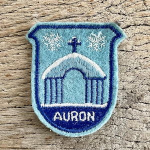 Op de afbeelding: Een lichtblauwe en marineblauwe geborduurde patch met het woord "AURON" onderaan. De patch heeft een gebouwontwerp met een kruis erop en sneeuwvlokken aan weerszijden. De patch ligt op een houten oppervlak.