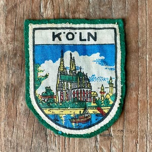 May include: A shield-shaped fabric patch featuring a printed illustration of the Cologne Cathedral and a boat on the water. The text K'O'LN is printed in black at the top. The patch has a white border and a dark green outer edge.