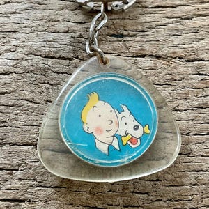 Pu&ograve; includere: Un portachiavi vintage con un'immagine a cartoni animati di un ragazzo con i capelli gialli e un cane bianco su sfondo blu. Il portachiavi ha una base in plastica trasparente triangolare e un portachiavi in metallo argentato.