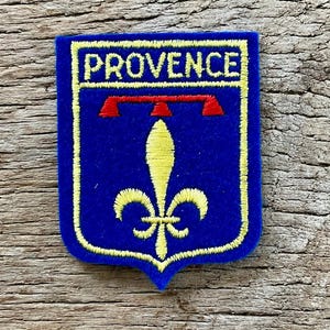 May include: A rectangular blue embroidered patch with rounded bottom corners featuring a yellow border and the word PROVENCE in yellow capital letters. Below the text is a red horizontal bar above a yellow fleur-de-lis emblem centered on the patch.