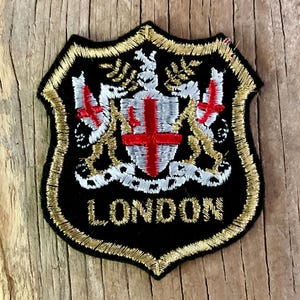 Op de afbeelding: Geborduurde patch met een schildontwerp, zwarte achtergrond en gouden rand. Het schild toont een wapen met een rood kruis en het woord "LONDON" in goud. De patch is ongeveer 6,35 cm breed.