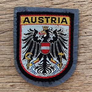 Peut inclure: Écusson rectangulaire avec le mot "AUSTRIA" au-dessus d'un emblème d'aigle noir avec un bouclier rouge et blanc, une couronne dorée et des détails dorés et bleus. Le patch a une bordure grise.