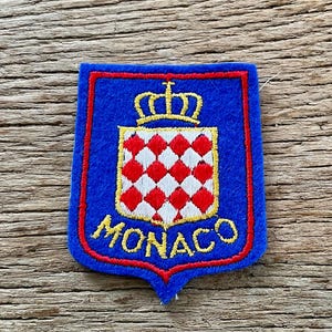 Op de afbeelding: Een blauwe geborduurde patch in de vorm van een schild met een rode rand. Het midden toont een gele kroon boven een wapenschild met een wit en rood ruitpatroon. Het woord MONACO is in gele hoofdletters onderaan de patch geborduurd.