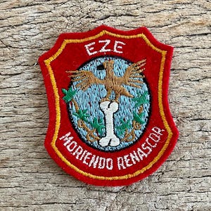 May include: Embroidered red patch with a gold border. The patch features a central design of a brown bird above a bone, set against a blue and green background. The word "EZE" is at the top, and "MORIENDO RENASCOR" is at the bottom.