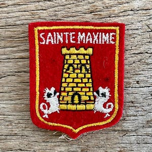 Op de afbeelding: Geborduurde patch met een rode achtergrond en een gele rand. De patch heeft een gele toren met de tekst "SAINTE MAXIME" in het wit. Twee witte zeepaardachtige wezens flankeren de toren. De patch is rechthoekig met een schildachtige vorm.