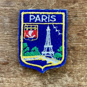 Puede incluir: Un parche bordado azul con un borde amarillo y la palabra "PARIS" en blanco. El parche presenta una imagen de la Torre Eiffel, árboles y un escudo de armas. El parche es probablemente un recuerdo o artículo de colección.