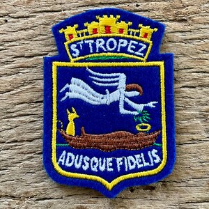 Op de afbeelding: Een blauwe geborduurde patch met een gele kroon aan de bovenkant, de tekst ST TROPEZ, en een centrale afbeelding van een witte engelfiguur die boven een bruine boot met een gele dierfiguur zweeft. De tekst ADUSQUE FIDELIS staat onderaan.