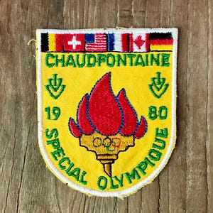 Op de afbeelding: Een gele geborduurde patch met een rode olympische fakkel en de olympische ringen aan de basis. De tekst luidt CHAUDFONTAINE 19 80 SPECIAL OLYMPIQUE. Boven de tekst staat een rij van zes nationale vlaggen. De patch heeft een witte rand.
