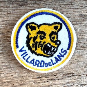 Op de afbeelding: Een ronde geborduurde patch met een gele en zwarte berenkopafbeelding gecentreerd op een witte achtergrond. De patch is omrand door een dunne blauwe en gele ring, met de blauwe tekst VILLARD DE LANS gebogen langs de onderrand.