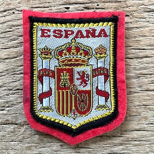 Op de afbeelding: Geborduurde patch met het woord "ESPANA" bovenaan. De patch toont het Spaanse wapen in het midden, met rode, gouden en witte details. De patch heeft een rode rand met zwarte en gouden afwerking.