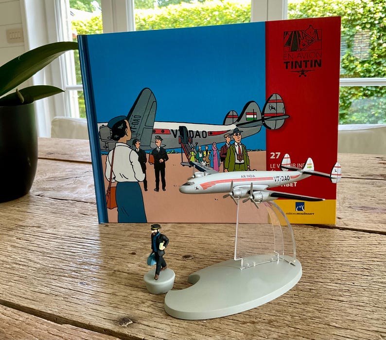 Op de afbeelding: Een modelvliegtuig met de tekst "AIR INDIA VT-DAD" wordt getoond voor een boek met een blauwe kaft en de tekst "EN AVION TINTIN". Er is ook een klein beeldje van een man in een zwart pak aanwezig.