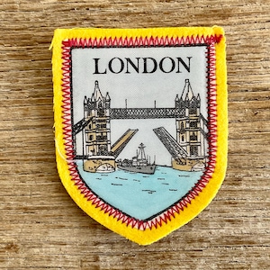 Op de afbeelding: Een schildvormige geborduurde patch met een felgele rand en rode stiksels. Het midden heeft een witte achtergrond met het woord LONDON in zwarte letters boven een illustratie van de Tower Bridge met een schip dat door de open brug vaart.