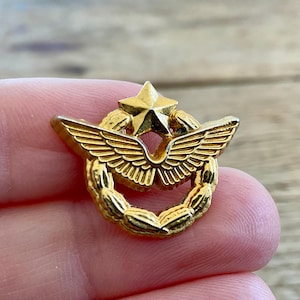 May include: A gold-colored military aviation badge featuring a five-pointed star at the top, wings in the center, and a laurel wreath encircling the wings. The badge is likely made of metal and has a shiny finish.