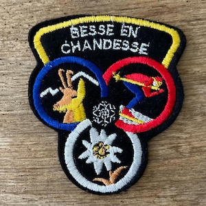May include: Embroidered patch with the text "BESSE EN CHANDESSE" in white letters on a black background. The patch features three interconnected circles with images of a skier, a flower, and a mountain goat. The patch is outlined in yellow.