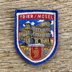 Puede incluir: Un parche bordado en forma de escudo que muestra la Porta Nigra de Tréveris, Alemania. El diseño incluye el texto TRIER / MOSEL y PORTA NIGRA, con un escudo central rojo que representa una figura sosteniendo una llave, flanqueado por dos uvas.