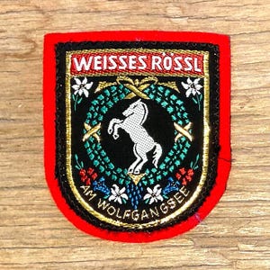 Puede incluir: Un parche rectangular con borde rojo y centro negro. El parche presenta el texto "WEISSES RÖSSL" en la parte superior y "AM WOLFGANGSEE" en la inferior. Un caballo blanco está en el centro, rodeado de una corona y adornos florales.