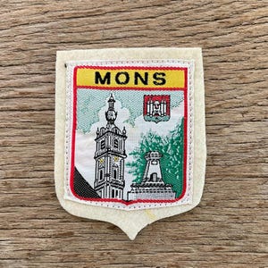 Pode incluir: Remendo de tecido em forma de escudo, com a palavra "MONS" em amarelo. O remendo apresenta uma ilustração detalhada de um edifício alto e uma torre, com detalhes em vermelho e verde. Possui uma borda vermelha e é costurado em um suporte creme, sobre uma superfície de madeira.