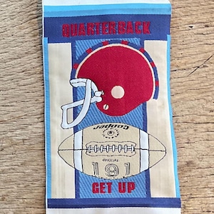 Op de afbeelding: Een rechthoekige stoffen patch met een voetbalthema. De bovenkant van de patch leest "QUARTERBACK" in rode letters. Hieronder een rode voetbalhelm en een voetbal met de tekst "OFFICIAL 191 GET UP". De achtergrond heeft blauwe en witte strepen.