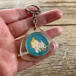 Pu&ograve; includere: Un portachiavi vintage con un'immagine a cartoni animati di un ragazzo e un cane su sfondo blu. Il portachiavi ha un ciondolo in plastica trasparente a forma di goccia e un portachiavi in metallo argentato.
