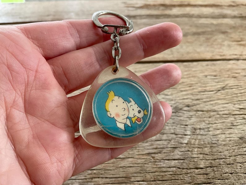 Pu&ograve; includere: Un portachiavi vintage con un'immagine a cartoni animati di un ragazzo e un cane su sfondo blu. Il portachiavi ha un ciondolo in plastica trasparente a forma di goccia e un portachiavi in metallo argentato.