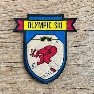 Op de afbeelding: Een vintage patch met een blauwe rand en een crèmekleurig midden met een skiër in rode kleding. De patch heeft de woorden "OLYMPIC SKI" in geel op een banner. De skiër draagt een skibril.