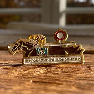 May include: A gold-toned pin featuring a horse race scene. The pin depicts a horse and jockey in motion, with a red and white racing marker. The text "HIPPODROME DE LONGCHAMP" is displayed on the pin.