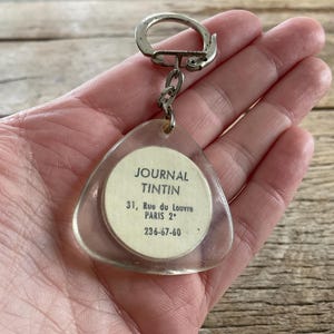 Pu&ograve; includere: Un portachiavi triangolare trasparente con anello e catena in metallo argentato. Il portachiavi presenta un inserto di carta circolare con il testo "JOURNAL TINTIN" e i dettagli dell'indirizzo. Lo sfondo &egrave; una superficie di legno.