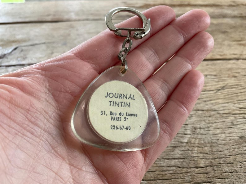 Pu&ograve; includere: Un portachiavi triangolare trasparente con anello e catena in metallo argentato. Il portachiavi presenta un inserto di carta circolare con il testo "JOURNAL TINTIN" e i dettagli dell'indirizzo. Lo sfondo &egrave; una superficie di legno.