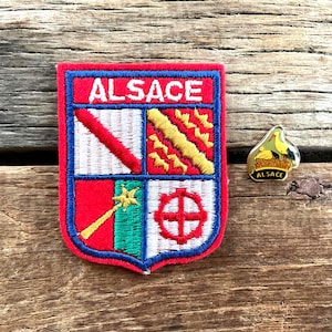 Op de afbeelding: Een rood en blauw geborduurd embleem met het woord "ALSACE" en een schildontwerp, naast een kleine speld met een ooievaar en het woord "ALSACE". Beide liggen op een houten oppervlak.