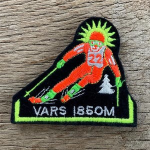 May include: Embroidered patch featuring a skier in orange and white racing attire, with the number 22. The skier is set against a black background with a green border, the text "VARS 1850M", and a stylized sun and tree.