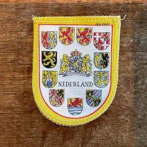 Pode incluir: Um patch com borda amarela com o brasão de armas dos Países Baixos. O patch tem um fundo branco com vários escudos heráldicos, um brasão central e a palavra "NEDERLAND" na parte inferior. O patch é provavelmente uma lembrança ou item de colecionador.