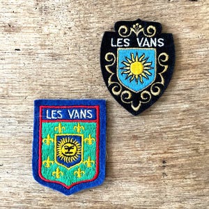 May include: Two embroidered patches with the text "LES VANS". One patch is black with a blue and yellow sun design. The other patch is blue, green, and red with a sun and fleur-de-lis design.