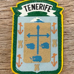 Puede incluir: Parche con la palabra "TENERIFE" en blanco sobre una pancarta verde. El parche tiene forma de escudo con borde dorado y contorno verde. El escudo tiene un centro beige con una cruz azul y formas azules, rodeado de anclas e iconos de castillo naranjas.