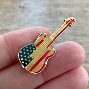 Puede incluir: Un pin en forma de guitarra con contorno dorado, que presenta el diseño de la bandera estadounidense. El pin tiene una sección azul con estrellas blancas y rayas rojas y blancas. El mástil de la guitarra es rojo con trastes dorados.