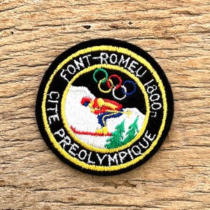 Puede incluir: Parche redondo negro con texto amarillo y un gráfico de esquiador. El parche dice "FONT-ROMEU 1800" y "CITE PREOLYMPIQUE". Los anillos olímpicos están encima del esquiador. El esquiador lleva ropa roja, amarilla y azul.
